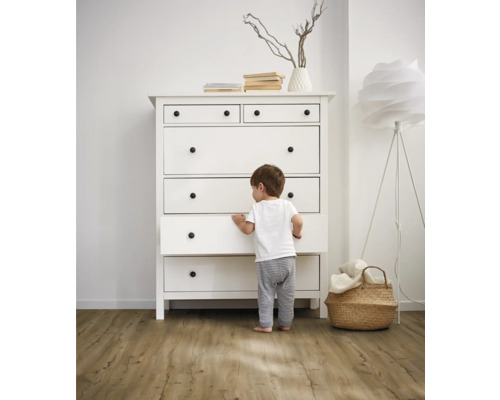 Dieťa stojí pred bielou komodou so zásuvkami a okrúhlymi úchytkami vo svetlej miestnosti.