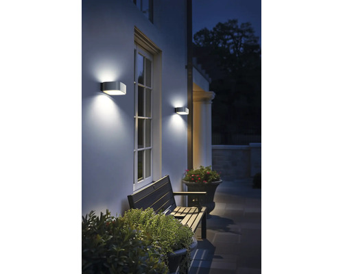 Vonkajšia scéna s domovou stenou, oknom, lavičkou a dvoma nástennými svietidlami.