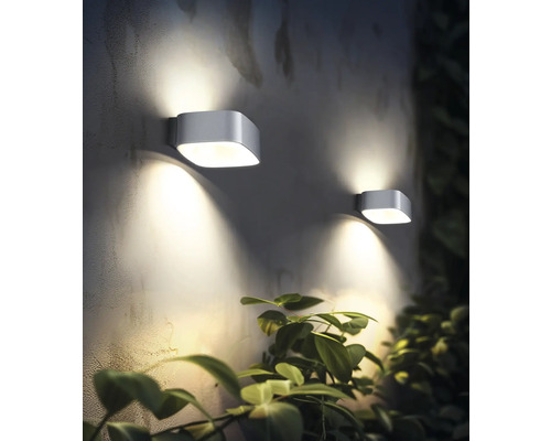 Dve moderné vonkajšie nástenné svietidlá osvetľujú stenu vedľa rastliny.