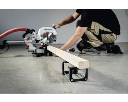 Osoba obsluhuje pokosovú pílu na rezanie kusa dreva, ktorý leží na podpernom stojane.