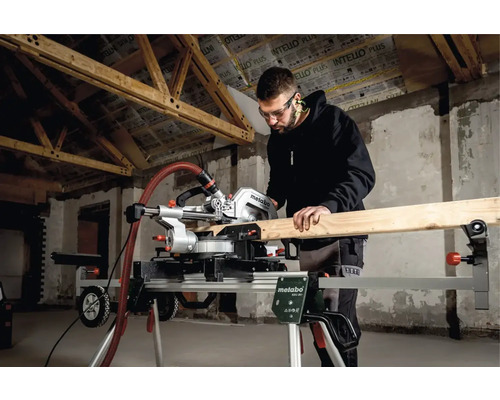 Muž s ochrannými okuliarmi reže drevo pokosovou pílou Metabo na podstavci v hrubej stavbe.