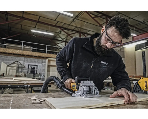 Muž používa drážkovaciu frézku na opracovanie obrobku. Náradie a drevo sa nachádzajú na pracovnom stole.