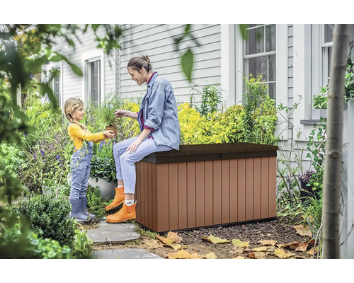 Záhradná scéna so ženou sediacou na hnedej záhradnej truhlici a malým dieťaťom, ktoré jej podáva rastlinu.