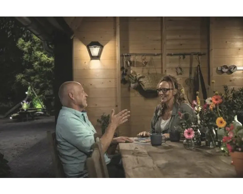 Pár sedí za dreveným stolom v jasne osvetlenom záhradnom domčeku s vonkajšou nástennou lampou.