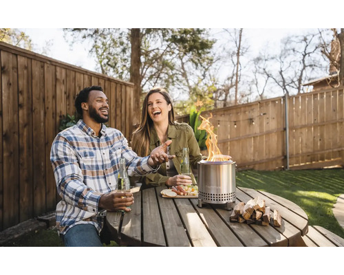 Vonkajšia scéna s párom pri drevenom stole s ohniskom, nápojmi a občerstvením.