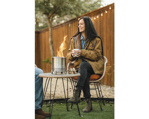Scéna so ženou sediacou pri záhradnom stole blízko ohniska a držiacej šálku.