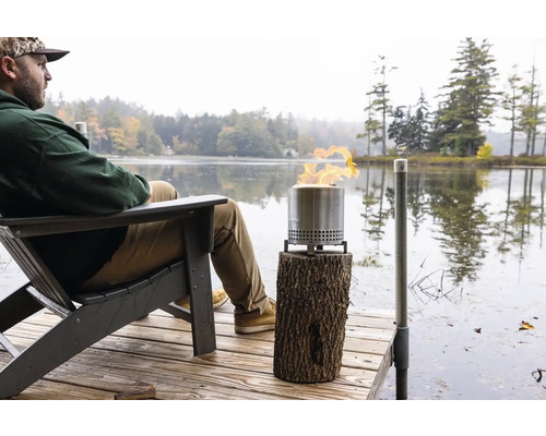 Scéna s mužom na drevenom móle pri jazere, vedľa neho horiace ohnisko na drevenej stoličke.