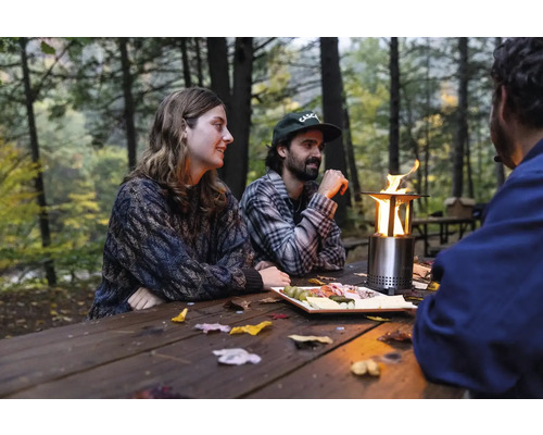 Skupina ľudí sedí vonku pri drevenom stole so stolovým ohniskom a jedlom