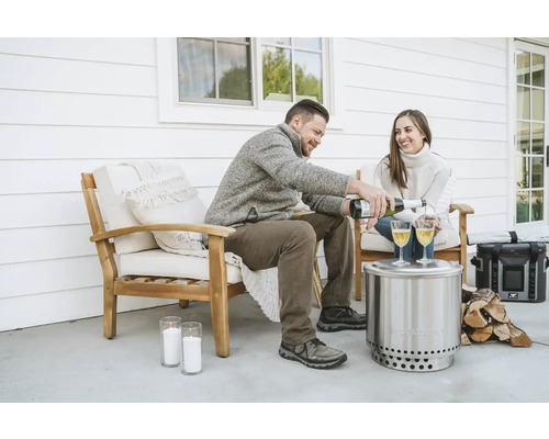 Scéna s párom sediacim vonku na stoličkách, zatiaľ čo muž nalieva sekt do pohárov na stole vedľa ohniska