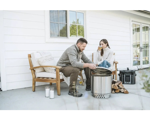 Scéna s mužom a ženou relaxujúcimi na terase, s nerezovým ohniskom bez dymu, palivovým drevom a nápojmi.