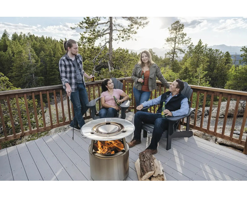 Scéna so štyrmi ľuďmi zhromaždenými okolo oceľového ohniska na terase so zábradlím, s kládami a stoličkami