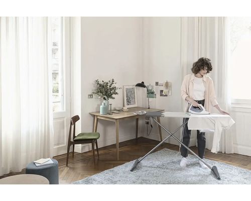 Svetlá izba so žehliacou doskou, žehličkou, písacím stolom a osobou pri žehlení