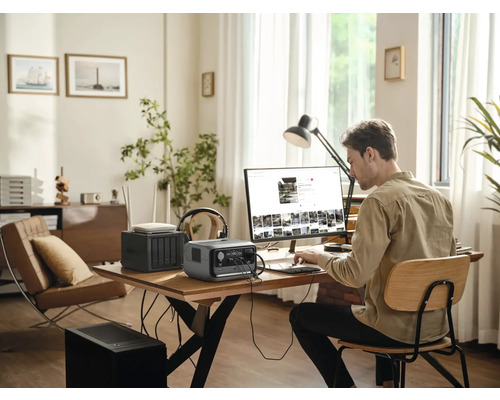 Muž pracuje na počítači s prenosnou elektrárňou, smerovačom a sieťovým úložným zariadením na stole v jasnej miestnosti.