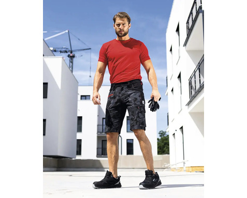 Muž na stavbe má na sebe červené tričko, krátke pracovné nohavice s maskovacím vzorom, bezpečnostnú obuv a rukavice