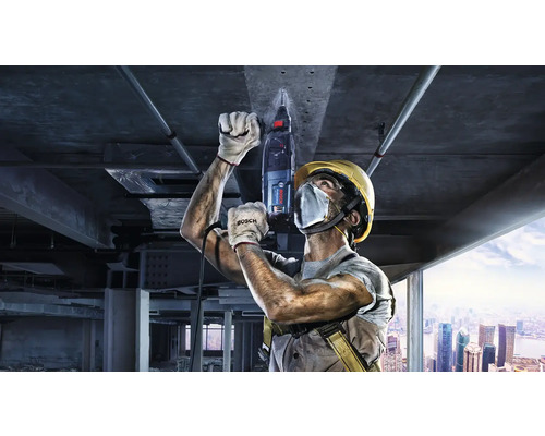 Stavebný robotník vŕta do stropu vŕtacím kladivom, v pozadí stavenisko.