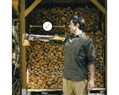 Muž drží štiepaciu sekeru Fiskars X25 v natiahnutej ruke pred hromadou dreva. Dôraz je kladený na správnu dĺžku ramena.