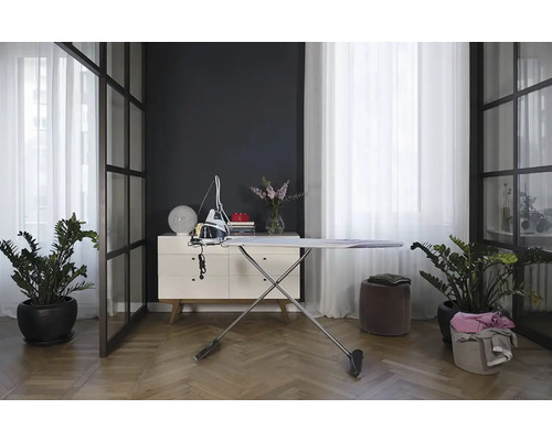 Žehlička a žehliaca doska v svetlej, modernej miestnosti s rastlinami a bielym príborníkom.