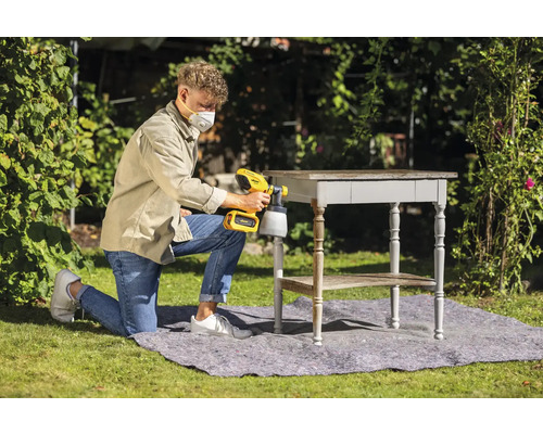 Muž používa vonku striekaciu pištoľ na lakovanie stola.