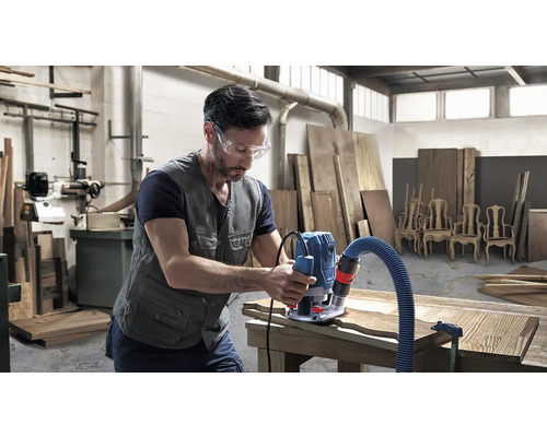 Muž s ochrannými okuliarmi opracováva drevo hornou frézou v dielni.