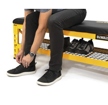 Osoba sedí na žltom pracovnom stole s úložným priestorom na topánky a zaväzuje si topánky. Na pracovnom stole sú ďalšie topánky. Logo DeWalt