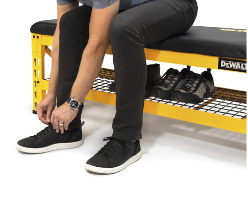 Osoba sedí na žltom pracovnom stole s úložným priestorom na topánky a zaväzuje si topánky. Na pracovnom stole sú ďalšie topánky. Logo DeWalt