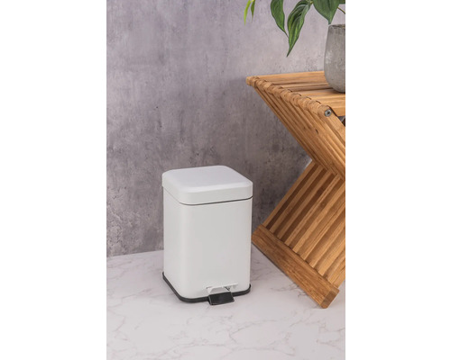 Biely kovový odpadkový kôš s pedálom vedľa dreveného odkladacieho stolíka