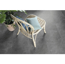 Scéna s ratanovým kreslom s vankúšmi a črepníkovou rastlinou na šedej podlahe.