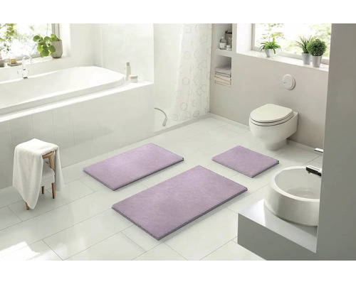 Svetlá kúpeľňa s vaňou, toaletou, umývadlom a súpravou troch fialových kúpeľňových predložiek.