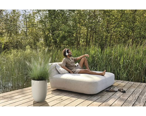 Muž relaxuje na záhradnom lehátku so slúchadlami, vedľa neho kvetináč s okrasnou trávou na drevenej terase.