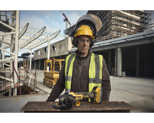 Stavebný robotník s ochrannou helmou a reflexnou vestou stojí pred staveniskom. Na stole leží akumulátorová uhlová brúska značky DeWalt.