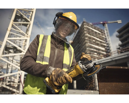 Stavebný robotník s ochrannou helmou, ochrannými okuliarmi a výstražnou vestou používa uhlovú brúsku na stavenisku.