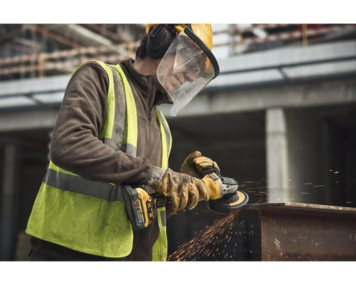 Stavebný robotník s ochrannými okuliarmi a žltou ochrannou prilbou obsluhuje uhlovú brúsku na kovovom nosníku. Lietajú iskry.