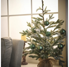 Dekoratívny vianočný stromček so zelenými guľami a jutovým vrecom na stole pred oknom.