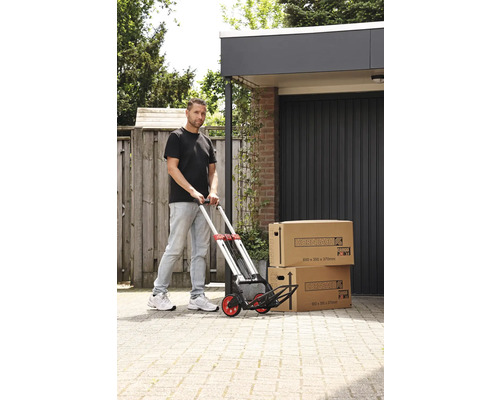 Muž tlačí ručný vozík s dvoma sťahovacími krabicami pred garážou.