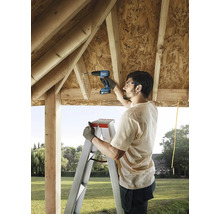 Muž používa akumulátorový skrutkovač na rebríku na stavbu dreveného domu.