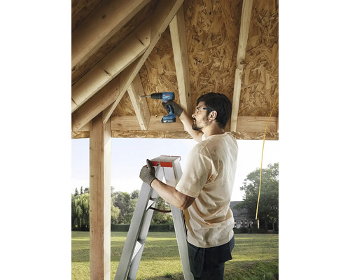 Muž používa akumulátorový skrutkovač na rebríku na stavbu dreveného domu.