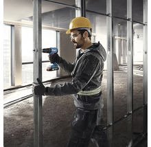 Stavebný robotník s ochrannou helmou a okuliarmi používa akumulátorový skrutkovač na kovovej konštrukcii.