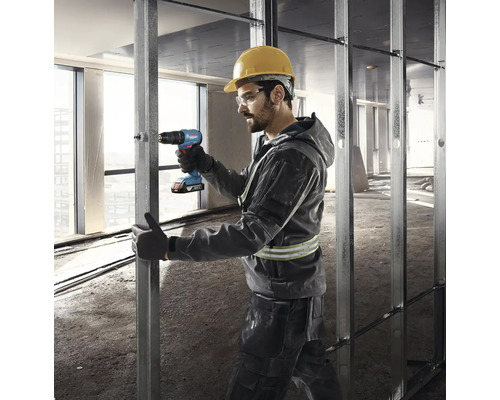 Stavebný robotník s ochrannou helmou a okuliarmi používa akumulátorový skrutkovač na kovovej konštrukcii.