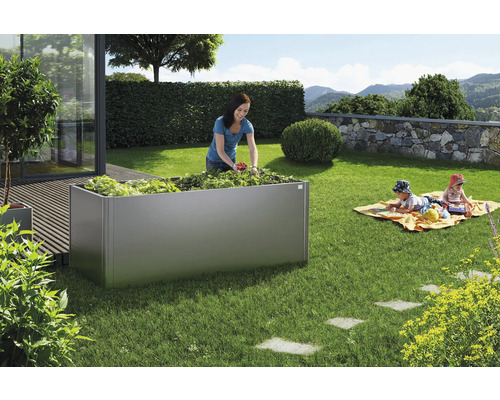 Záhradná scéna s vyvýšeným kovovým záhonom, ženou zbierajúcou úrodu a hrajúcimi sa deťmi