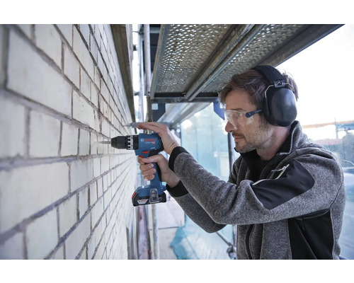 Muž vŕta akumulátorovou vŕtačkou Bosch do tehlovej steny, má ochranné okuliare a chrániče sluchu.