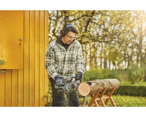 Muž pília kus dreva na kozlíku pomocou akumulátorovej reťazovej píly, pričom má ochranné okuliare a slúchadlá.