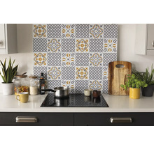 Organizovaná svetlá kuchyňa so zadnou stenou so sivo-žltým vzorom dlaždíc nad varnou doskou na bielej pracovnej doske s doplnkami a rastlinami.