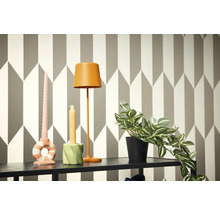 Dekoratívna scéna s lampou, sviečkami a črepníkovou rastlinou na poličke pred geometrickou tapetou.