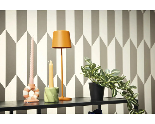 Dekoratívna scéna s lampou, sviečkami a črepníkovou rastlinou na poličke pred geometrickou tapetou.