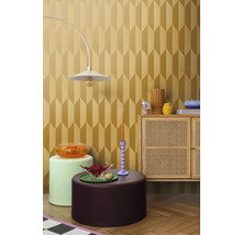 Ukážka vybavenia interiéru s lampou, rôznymi stolíkmi a skrinkou pred tapetou s geometrickým vzorom.