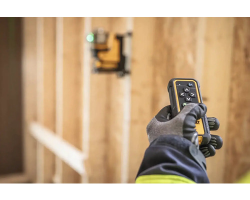 Ruka v rukavici drží diaľkové ovládanie lasera DeWalt pred drevenou konštrukciou.