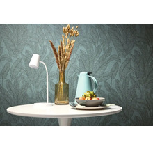 Stôl s lampou, vázou, termoskou a misou pred tapetou s listovým vzorom.