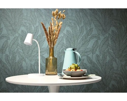 Stôl s lampou, vázou, termoskou a misou pred tapetou s listovým vzorom.