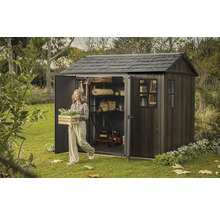 Záhradná scéna s domčekom na náradie, kosačkou na trávu a ženou nesúcou debničku s kvetmi.