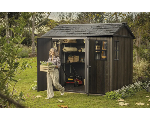 Záhradná scéna s domčekom na náradie, kosačkou na trávu a ženou nesúcou debničku s kvetmi.